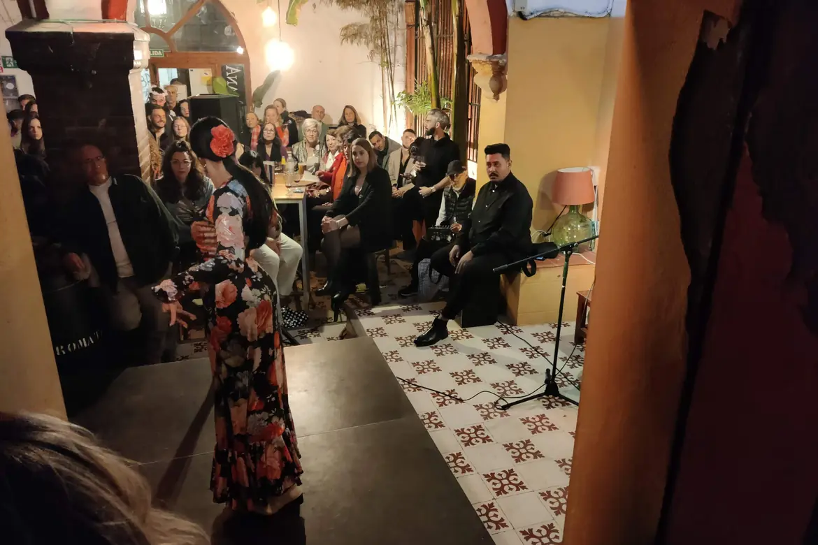 A dancer in a flowered dress performs on a small stage while a crowded courtyard audience watches from tables
