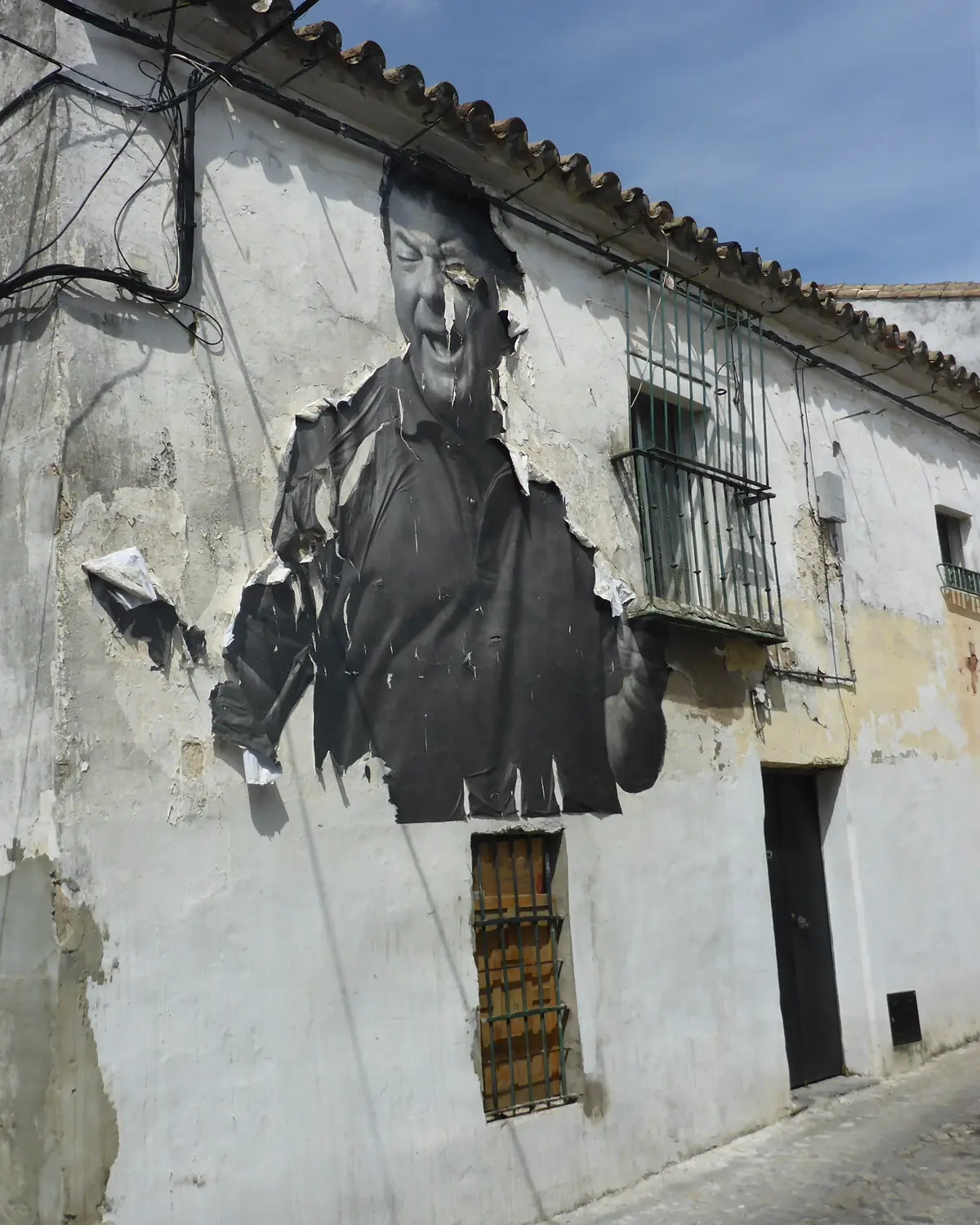 A large black-and-white portrait of a laughing flamenco singer pasted on a peeling whitewashed house wall in Jerez, with a barred window beside it