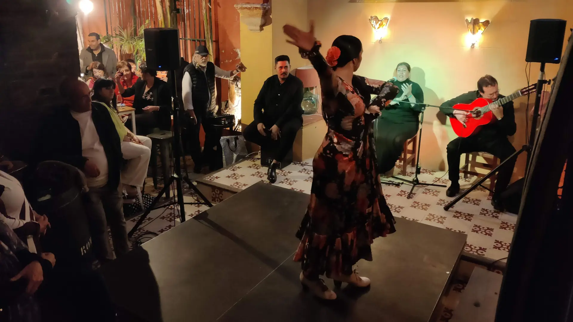 A flamenco dancer in a flowered dress raises one arm in front of a seated singer and a guitarist playing a red guitar, with an audience watching from the left